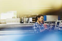 Female Asian factory worker operating a touch-screen display. Hair in a ponytail. Checkered shirt. Primary color blue.